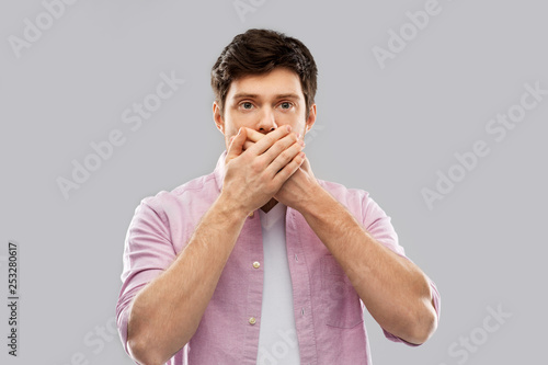 emotion, expression and people concept - shocked and speechless young man covering his mouth by hands over grey background