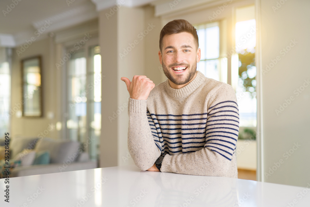 Young handsome man at home smiling with happy face looking and pointing to the side with thumb up. Young handsome man at home smiling with happy face looking and pointing to the side with thumb up.