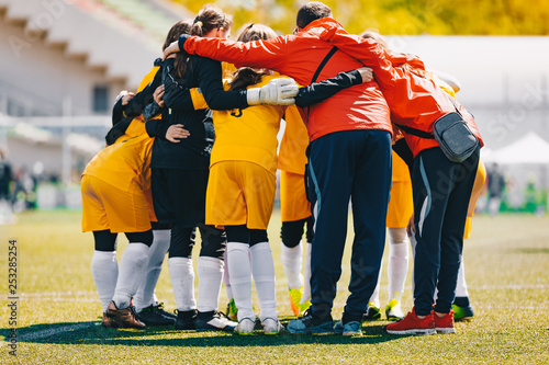Fototapeta Naklejka Na Ścianę i Meble -  Coach coaching Girls Sports Team. Girls school sports team huddling with coach on the grass field. Soccer football junior girls team at sports outdoor field before match. Coach Building Confidence