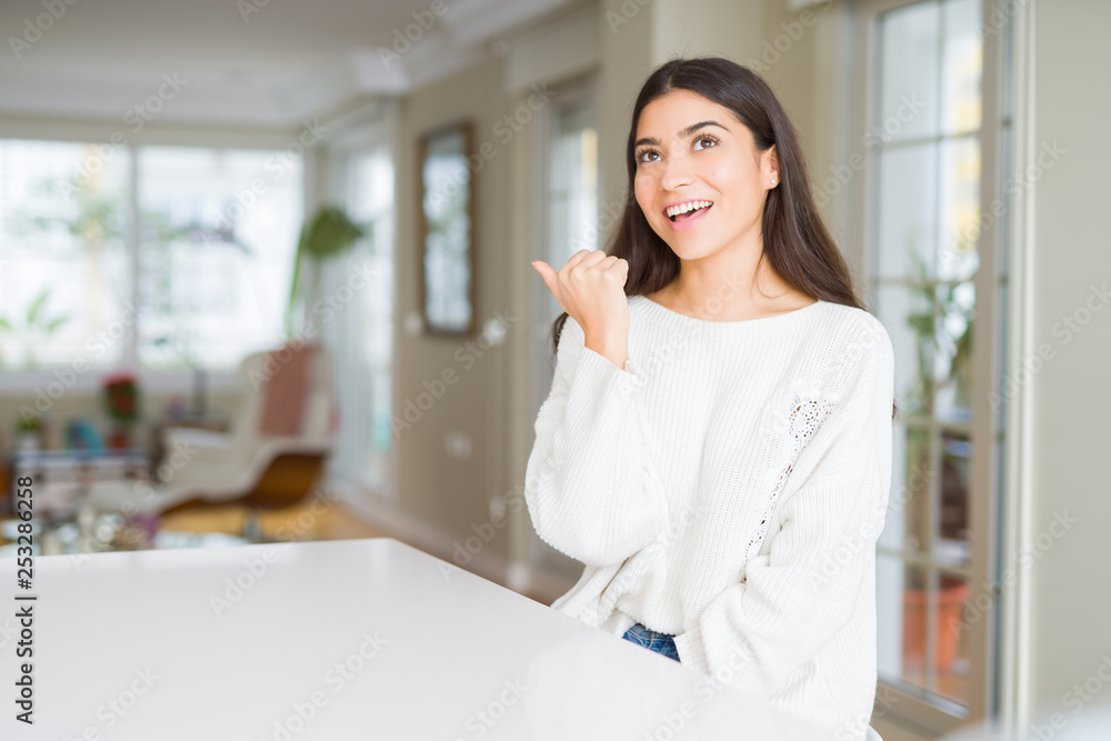 Young beautiful woman at home on white table smiling with happy face looking and pointing to the side with thumb up.