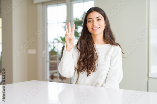 Young beautiful woman at home on white table showing and pointing up with fingers number three while smiling confident and happy.