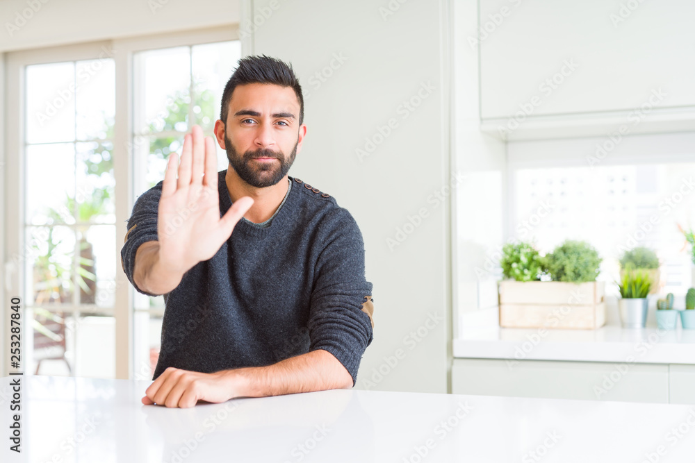 Handsome hispanic man wearing casual sweater at home doing stop sing with palm of the hand. Warning expression with negative and serious gesture on the face.