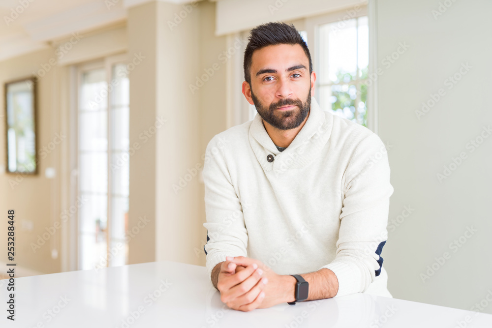 Handsome hispanic man wearing casual white sweater at home Relaxed with serious expression on face. Simple and natural looking at the camera.