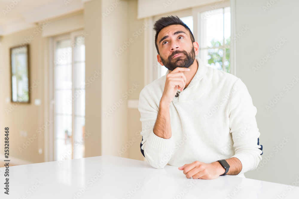 Handsome hispanic man wearing casual white sweater at home with hand on chin thinking about question, pensive expression. Smiling with thoughtful face. Doubt concept.