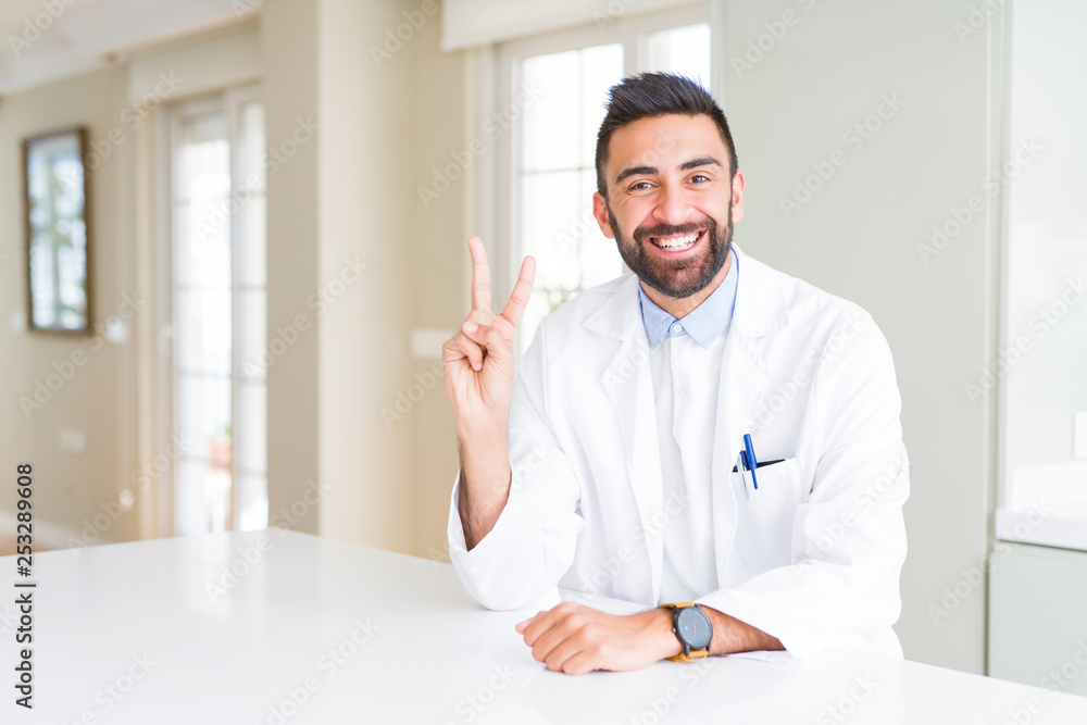 Handsome hispanic doctor or therapist man wearing medical coat at the clinic showing and pointing up with fingers number two while smiling confident and happy.
