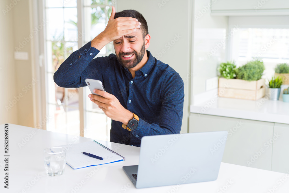 Handsome hispanic business man using smartphone and laptop at the office stressed with hand on head, shocked with shame and surprise face, angry and frustrated. Fear and upset for mistake.