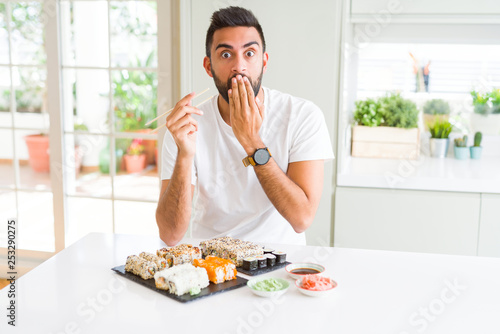 Handsome hispanic man eating asian sushi using chopsticks cover mouth with hand shocked with shame for mistake, expression of fear, scared in silence, secret concept