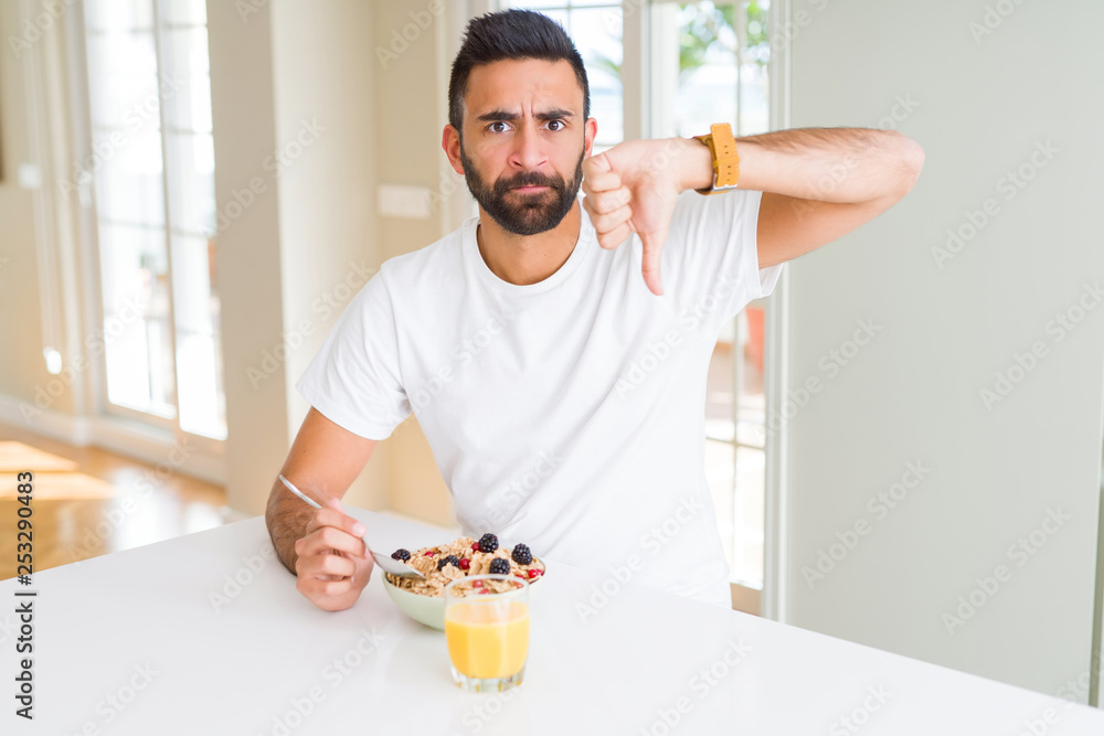 Handsome hispanic man eating healthy breakfast in the morning at home with angry face, negative sign showing dislike with thumbs down, rejection concept Handsome hispanic man eating healthy breakfast in the morning at home with angry face, negative sign showing dislike with thumbs down, rejection concept