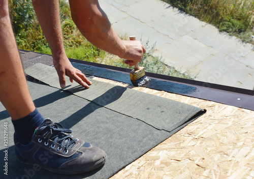 Tableau sur toile Roofer contractor gluing waterproof membrane on wooden roof top surface with brush and black bitumen  spray on tar before installing asphalt shingles