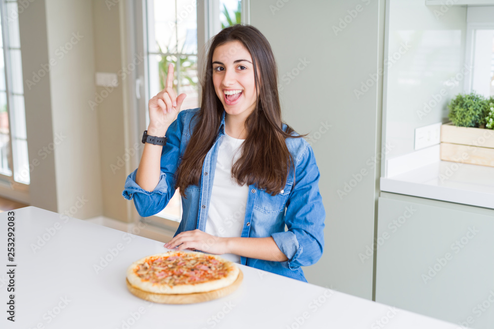 Beautiful young woman eating homemade tasty pizza at the kitchen pointing finger up with successful idea. Exited and happy. Number one. Beautiful young woman eating homemade tasty pizza at the kitchen pointing finger up with successful idea. Exited and happy. Number one.
