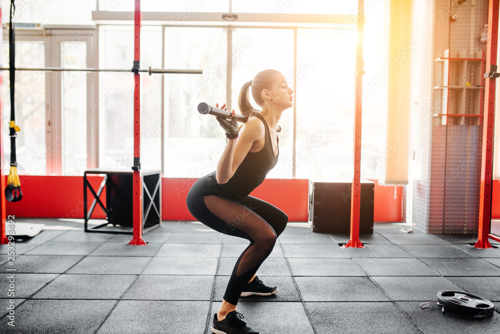Obraz premium Beautiful brunette during workout in gym. Sporty girl in black sportswear squats with barbell training. Side view
