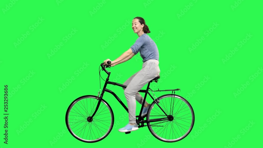 Smiling woman riding a bicycle over a green screen, rising her legs to the chassis, looking upwards, waving hello. No motion blur for optimal keying. Smiling woman riding a bicycle over a green screen, rising her legs to the chassis, looking upwards, waving hello. No motion blur for optimal keying.