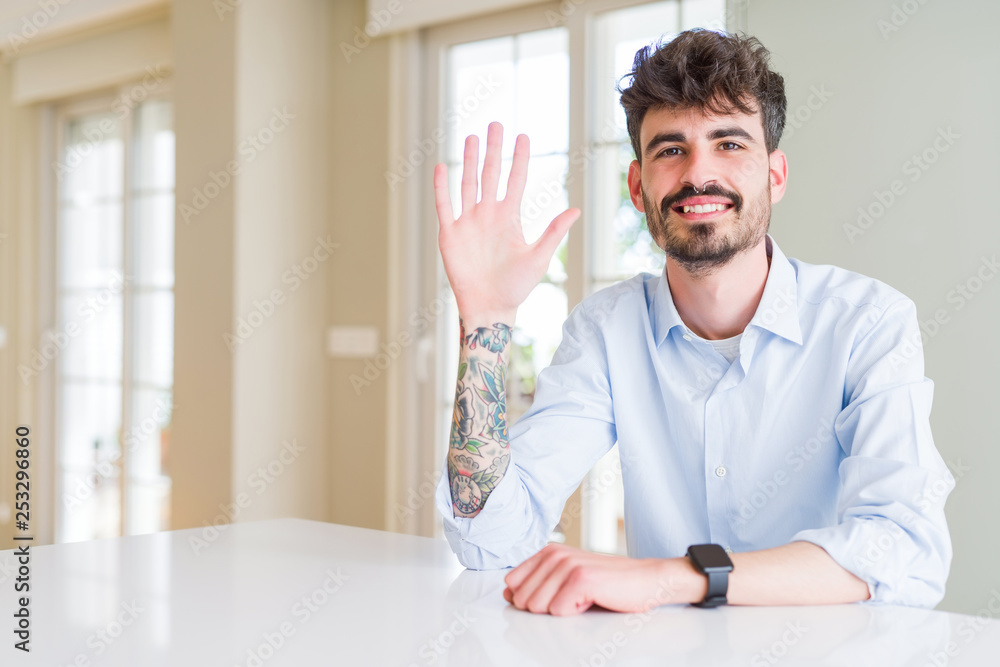 Young businesss man sitting on white table showing and pointing up with fingers number five while smiling confident and happy.