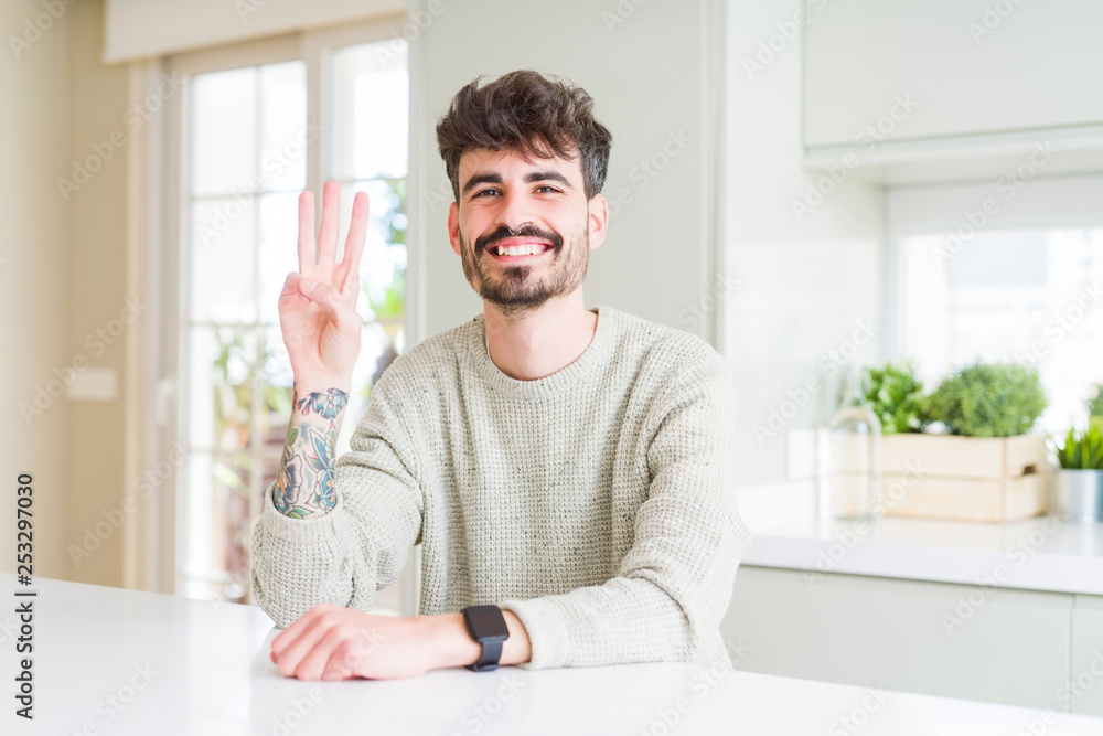 Young man wearing casual sweater sitting on white table showing and pointing up with fingers number three while smiling confident and happy. Young man wearing casual sweater sitting on white table showing and pointing up with fingers number three while smiling confident and happy.