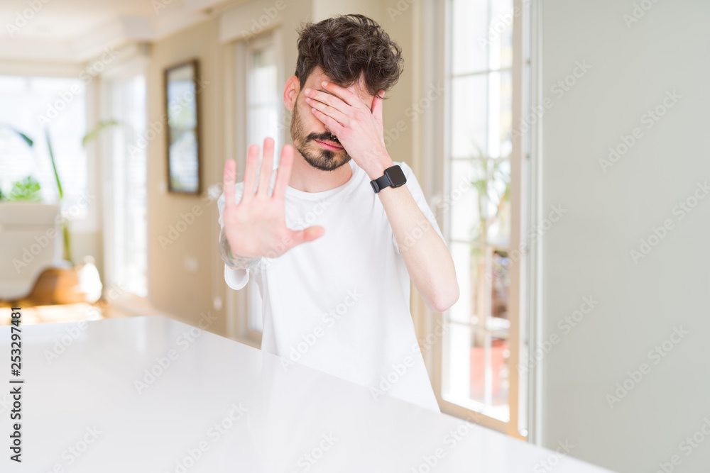 Young man wearing casual t-shirt sitting on white table covering eyes with hands and doing stop gesture with sad and fear expression. Embarrassed and negative concept.