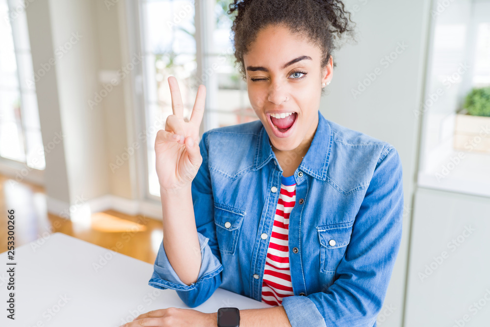 Beautiful young african american woman with afro hair wearing casual denim jacket smiling with happy face winking at the camera doing victory sign. Number two.