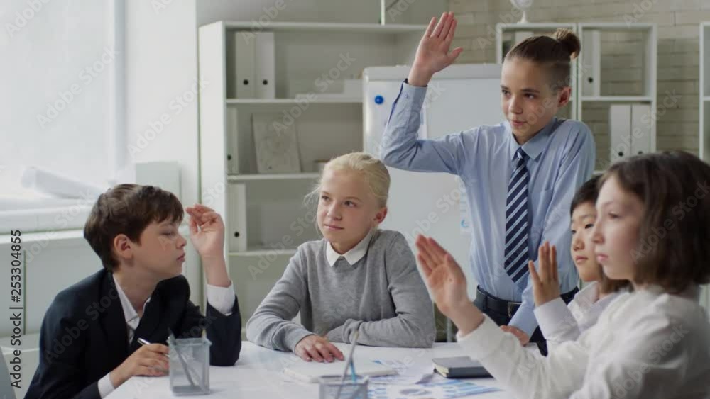 Panning of several school kids gathered at desk and rising their hands to answer teachers question Panning of several school kids gathered at desk and rising their hands to answer teachers question