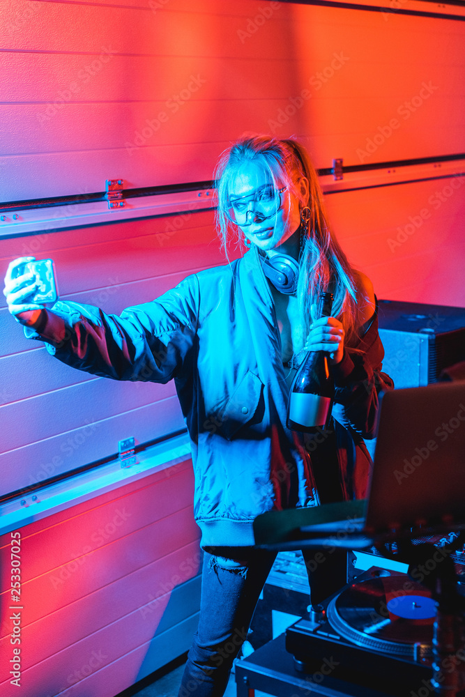 stylish blonde dj woman holding bottle and taking selfie in nightclub
