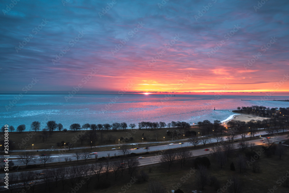A beautiful sunrise landscape photograph of the bright orange and yellow sun peering over the horizon on Lake Michigan in Chicago with pink and blue clouds in the sky and cars on Lake Shore Drive.
