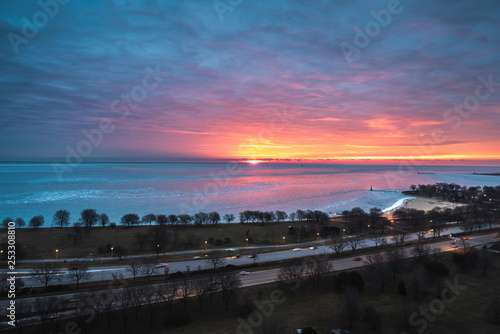 A beautiful sunrise landscape photograph of the bright orange and yellow sun peering over the horizon on Lake Michigan in Chicago with pink and blue clouds in the sky and cars on Lake Shore Drive.