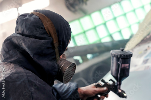 Man with protective clothes and mask painting car using spray compressor.