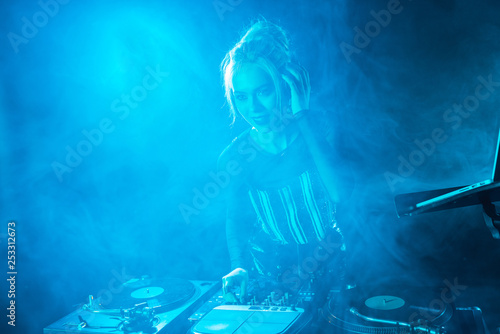 cheerful blonde dj girl listening music in headphones near dj equipment in nightclub with smoke