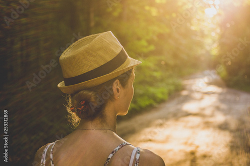 A woman in a straw hat on a street in summer looks thoughtfully at the road ahead. View from the back