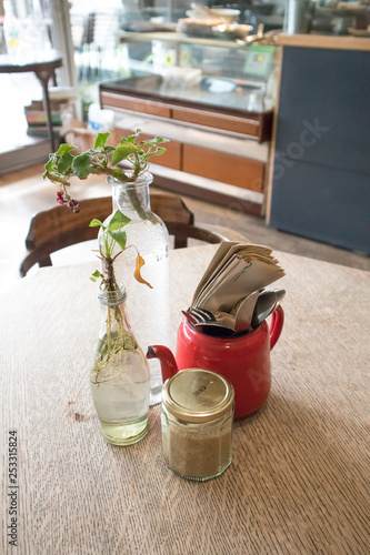 Interior shot of cafe table with homemade centre decorations.  Glass bottles with plants, sygar in jar and teapot with cutelry and napkins
