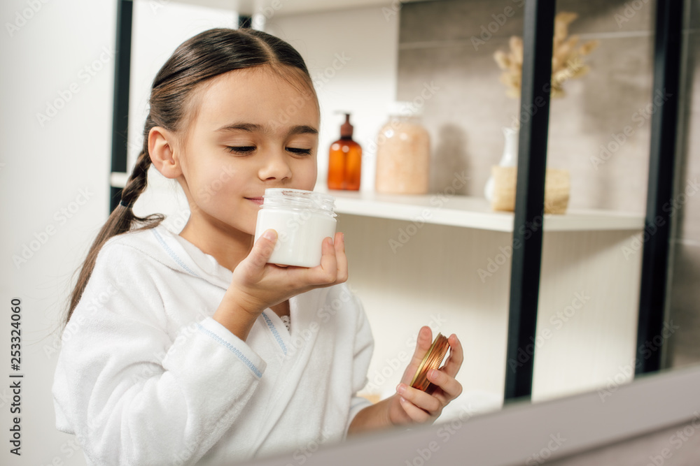 selective focus of child in white bathrobe looking to mirror and sniffing cosmetic cream in bathroom selective focus of child in white bathrobe looking to mirror and sniffing cosmetic cream in bathroom