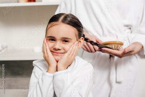 mother in white bathrobe combing daughter hair with wooden hairbrush in bathroom