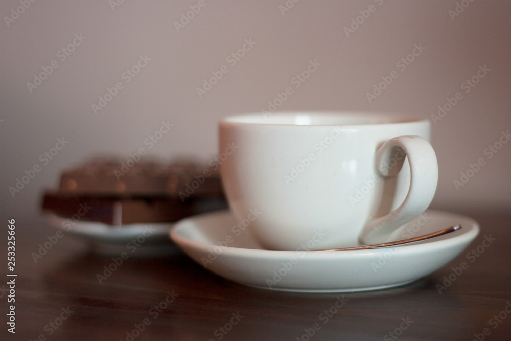 Obraz premium White cup on a saucer with metal spoon with chocolate biscuits in the background. Coffee shop, tea shop, cuppa with friends, relaxing, take a break concepts.