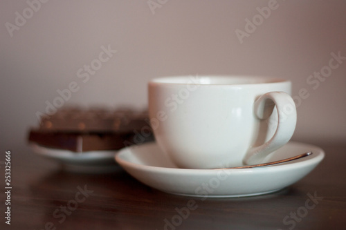 White cup on a saucer with metal spoon with chocolate biscuits in the background. Coffee shop, tea shop, cuppa with friends, relaxing, take a break concepts.