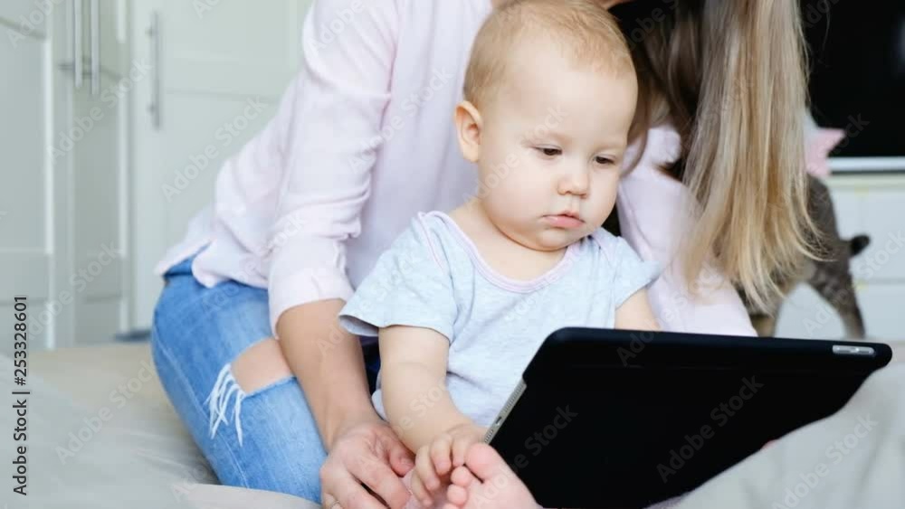 Little cute baby is looking at the tablet with mom in the cozy bedroom on the background of the cat. Concept of development and education of children Little cute baby is looking at the tablet with mom in the cozy bedroom on the background of the cat. Concept of development and education of children