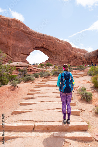 Young woman wearing backpack is looking at the North Window at Arches National Park in Utah. Female hiker in Arches National Park. Travel and adventure concept.