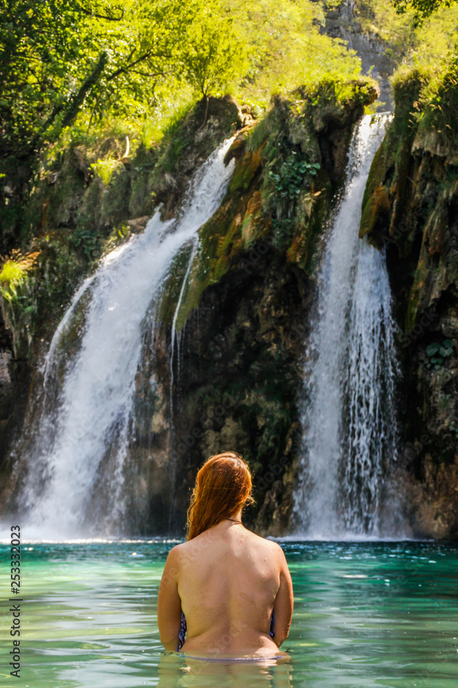 Young plump red-haired white woman with a bare back is standing in the turquoise water of a beautiful waterfall among the rocks in the jungle Young plump red-haired white woman with a bare back is standing in the turquoise water of a beautiful waterfall among the rocks in the jungle