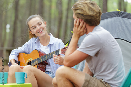 young couple sat by tent woman playing guitar