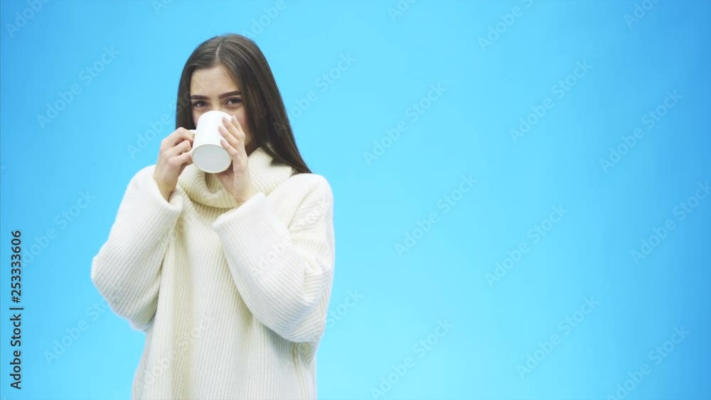Interestingly, a young woman in a knitted white sweater. Holds a white cup of coffee or tea in his hands. Isolated against the background of a blue wall, studio portrait. Concept of a way of life of Interestingly, a young woman in a knitted white sweater. Holds a white cup of coffee or tea in his hands. Isolated against the background of a blue wall, studio portrait. Concept of a way of life of
