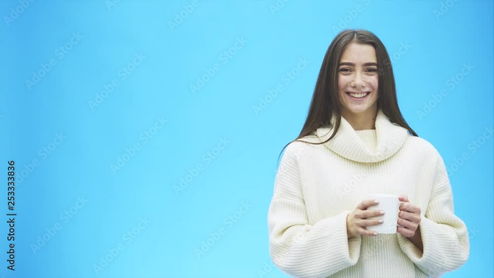 Interestingly, a young woman in a knitted white sweater. Holds a white cup of coffee or tea in his hands. Isolated against the background of a blue wall, studio portrait. Concept of a way of life of Interestingly, a young woman in a knitted white sweater. Holds a white cup of coffee or tea in his hands. Isolated against the background of a blue wall, studio portrait. Concept of a way of life of