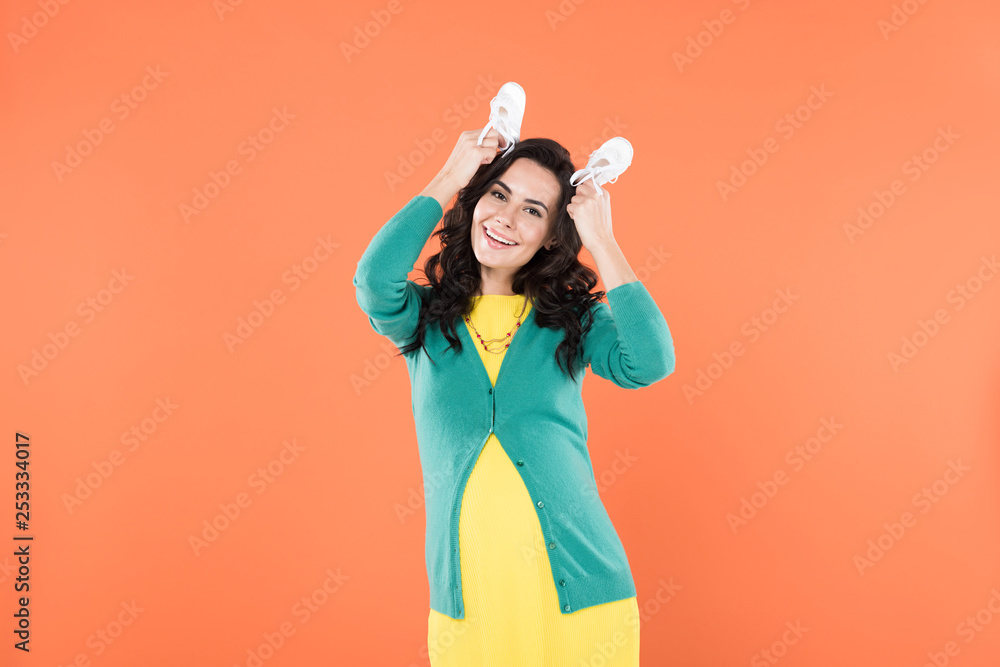 Carefree pregnant woman holding baby shoes isolated on orange Carefree pregnant woman holding baby shoes isolated on orange