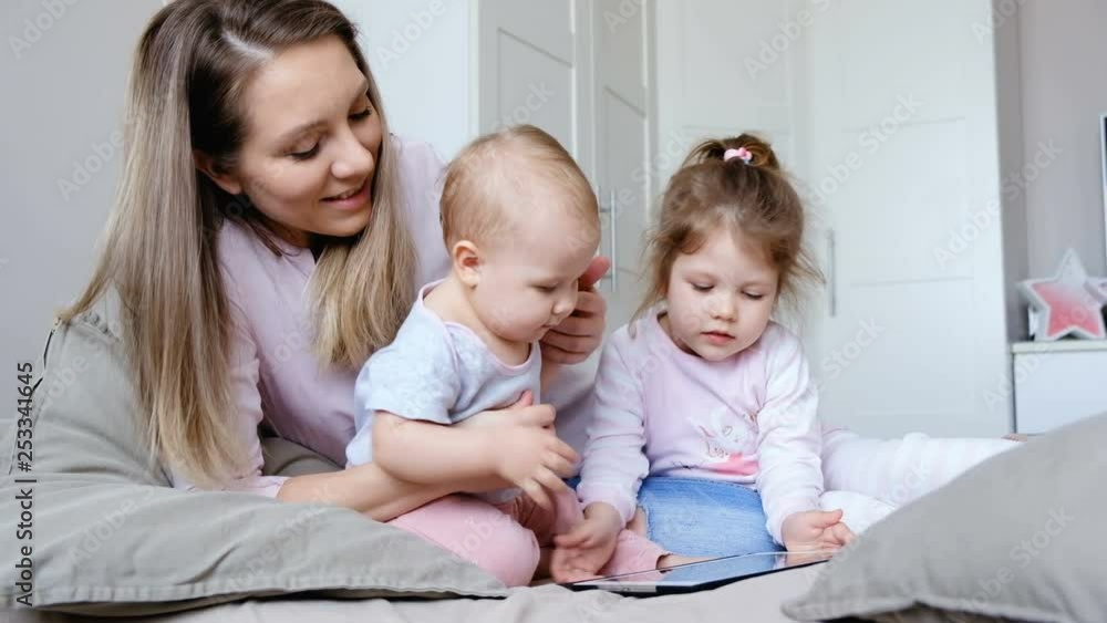 Cute young mother and her little son with a daughter are watching a cheerful cartoon on a tablet sitting in the bedroom on a weekend. Concept of family happiness Cute young mother and her little son with a daughter are watching a cheerful cartoon on a tablet sitting in the bedroom on a weekend. Concept of family happiness