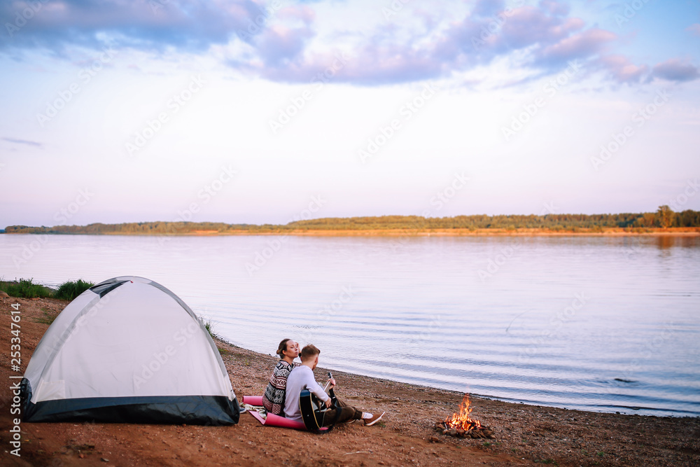 A man and a girl sit by the river and campfire and play the guitar.