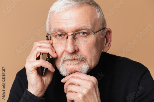 Old handsome man with gray hair and beard in eyeglasses and sweater thoughtfully looking in camera while talking on cellphone over beige background