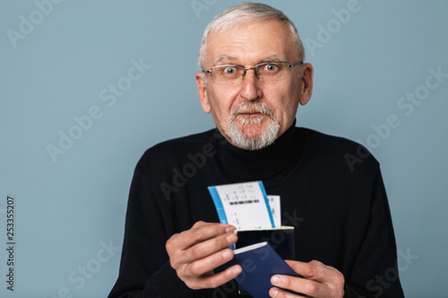 Old attractive man with gray hair and beard in eyeglasses and sweater holding tickets and passports in hands while amazedly looking in camera over blue background