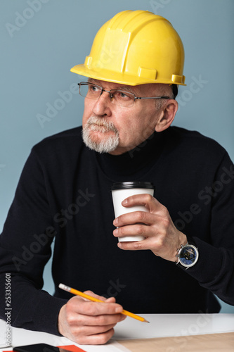 Old attractive man with gray beard in eyeglasses and yellow safety helmet holding cup of coffee to go in hand while thoughtfully looking aside over blue background
