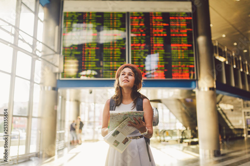 theme travel and transportation. Beautiful young caucasian woman in dress and backpack standing inside train station terminal looking at electronic scoreboard holding phone, map paper hand navigation photo