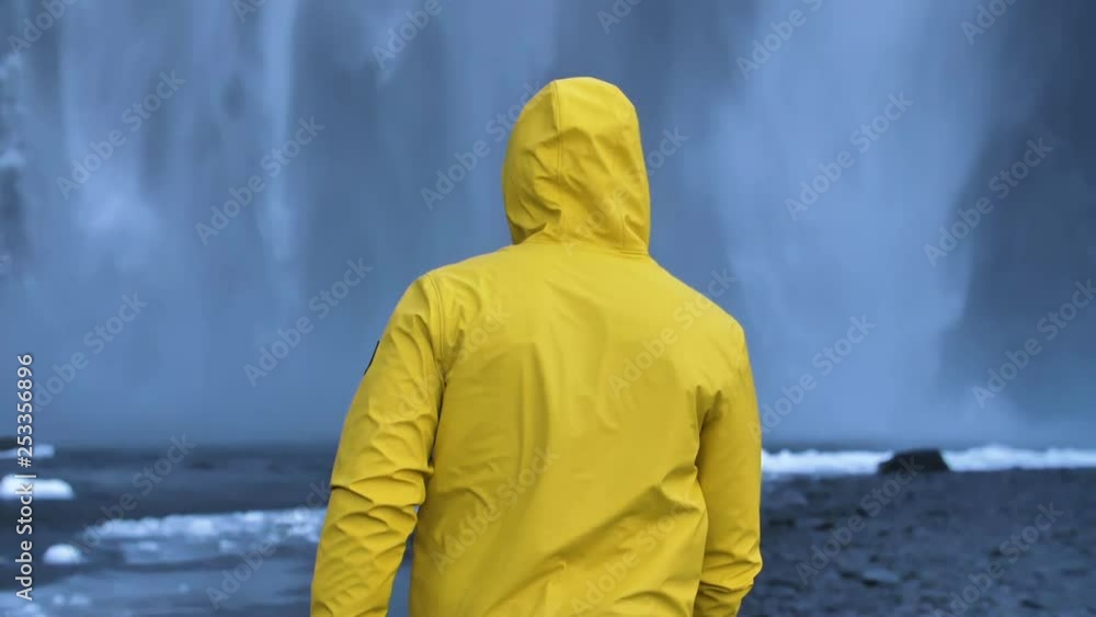 Young Male Traveler in yellow rain coat in front of huge frozen waterfall in slow motion Young Male Traveler in yellow rain coat in front of huge frozen waterfall in slow motion