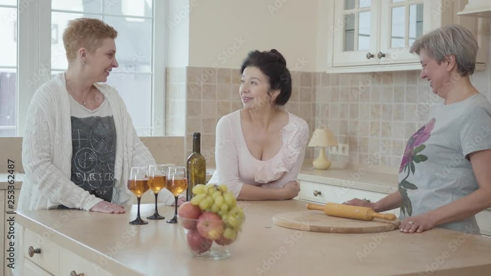 Elderly girlfriends have not seen each other for a long time and tell interesting stories. Adult girlfriends laugh and have fun at the table with fruit and wine in the kitchen. Elderly girlfriends have not seen each other for a long time and tell interesting stories. Adult girlfriends laugh and have fun at the table with fruit and wine in the kitchen.