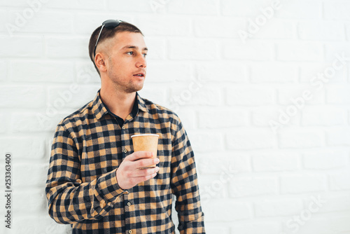 A handsome man in a plaid shirt on a brick background drinks coffee from a paper Cup