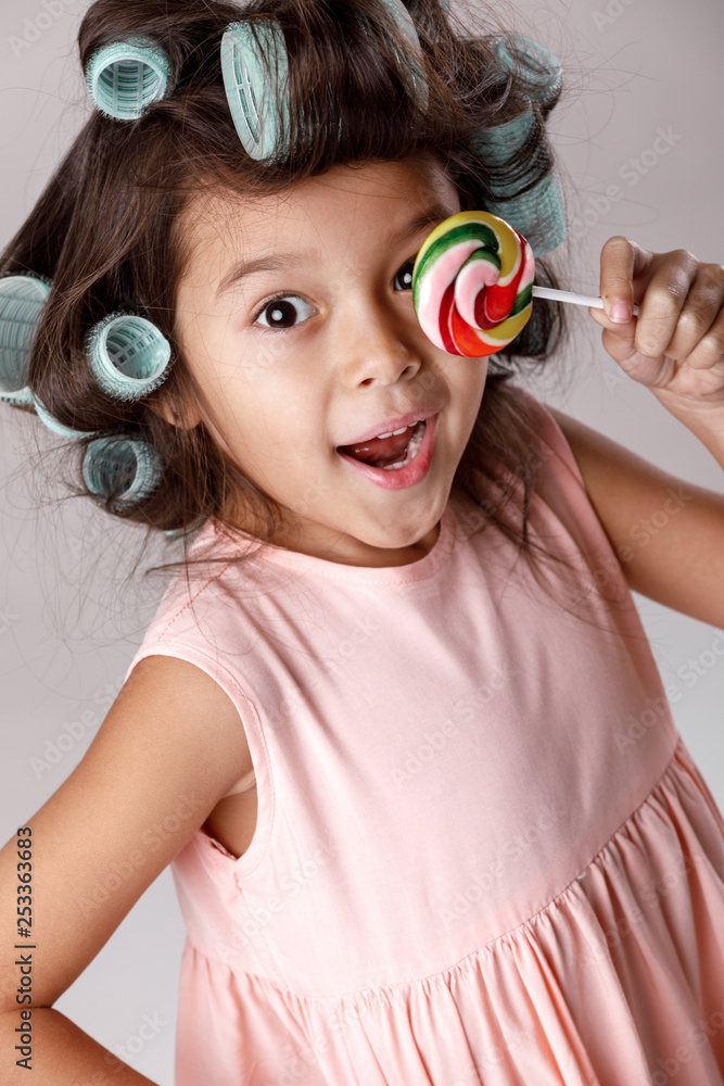 Φωτογραφία Funny little child girl in pink dress and hair curlers holding lollipop on gray background