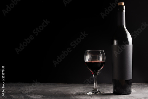 A bottle of red expensive wine with a black blank label on a dark background with a glass of red wine. Wine bottle mockup with space for text.